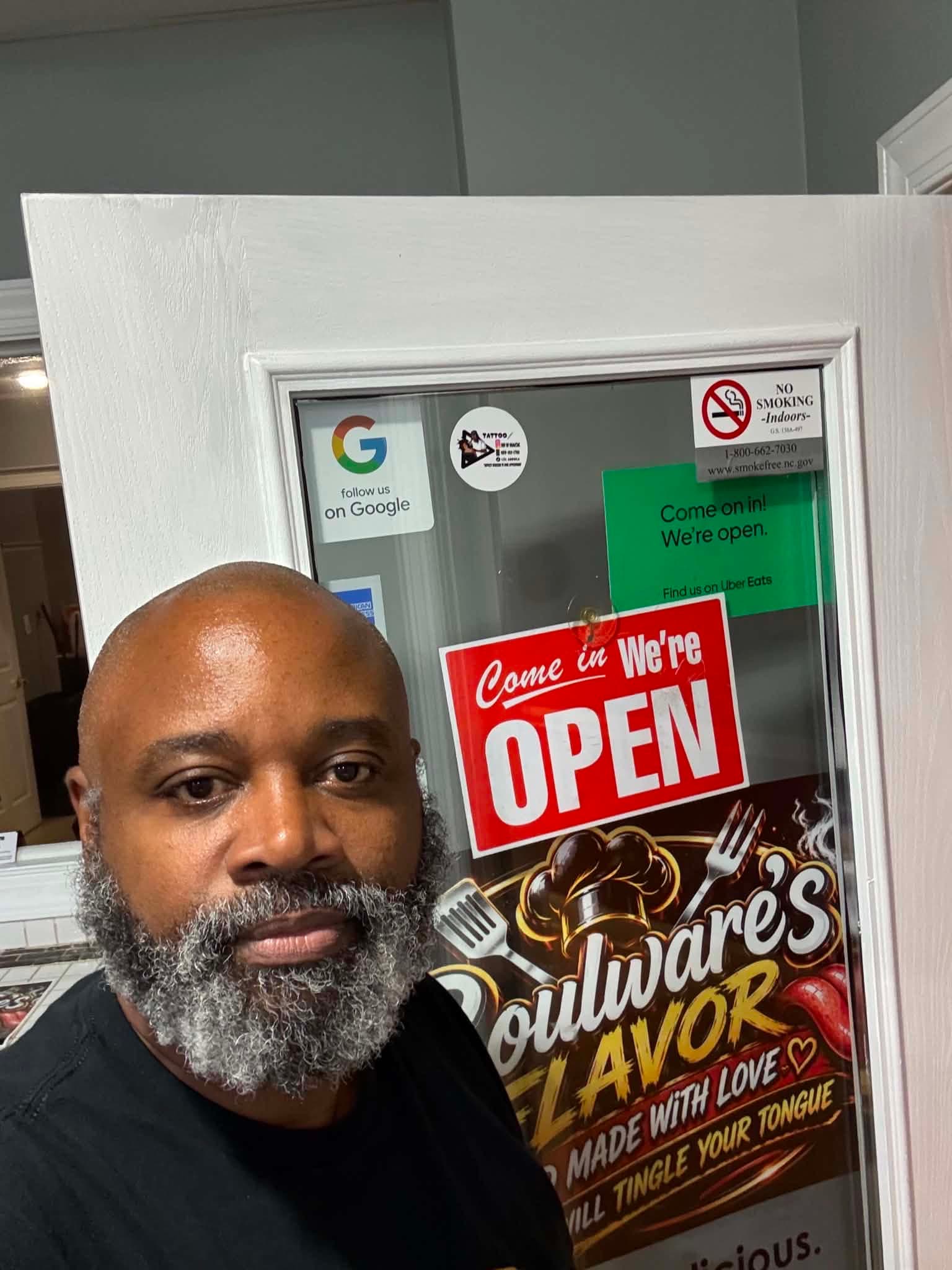 The owner of Boulware's Flavor Café at the café's front door, beside the Open sign and the Boulware's Flavor poster